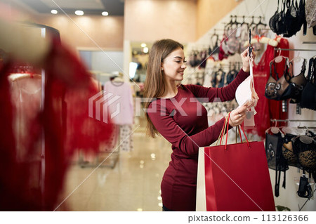 Young woman chooses sexy brassiere among set in a boutique. Pretty girl considers bra with shopping bags in her hand in underwear store. 113126366