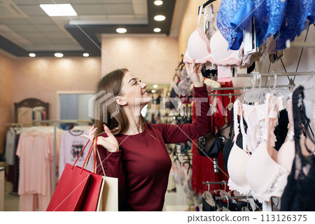 Young woman chooses sexy brassiere among set in a boutique. Pretty girl considers bra with shopping bags in her hand in underwear store. Young woman chooses sexy brassiere among set in a boutique. Pretty girl considers bra with shopping bags in her hand in underwear store. 113126375