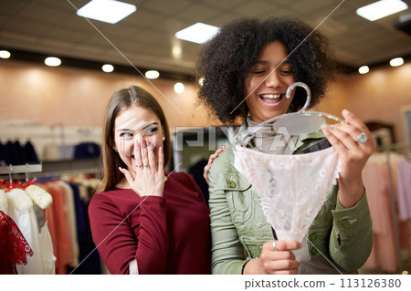 Two happy chic young mixed race woman shopping for lingerie in a clothing boutique with one holding panties as they decide whether to purchase. African american girl considers underwear with friend. 113126380