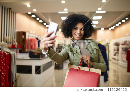 Cute african american woman taking selfie with shopping bags and smiling near clothing store. Black pretty girl taking photo on smartphone or talking on video call chat after visiting mall sale. 113126413