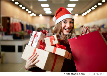 Happy girl shopping gifts in mall on christmas sale. New year holidays shopping idea concept. Smiling woman with colorful paper presents bags and gift boxes wearing christmas hat in store or shop. 113126448
