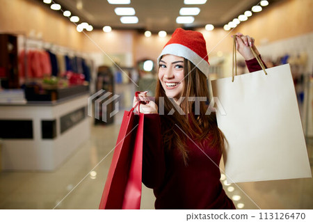Happy girl shopping gifts in mall on christmas sale. New year holidays shopping idea concept. Smiling pretty caucasian woman with colorful paper presents bags wearing christmas hat in store or shop. Happy girl shopping gifts in mall on christmas sale. New year holidays shopping idea concept. Smiling pretty caucasian woman with colorful paper presents bags wearing christmas hat in store or shop. 113126470