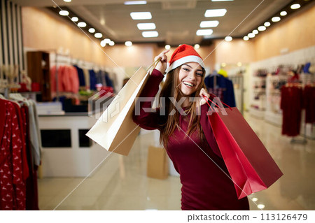 Happy girl shopping gifts in mall on christmas sale. New year holidays shopping idea concept. Smiling pretty caucasian woman with colorful paper presents bags wearing christmas hat in store or shop. Happy girl shopping gifts in mall on christmas sale. New year holidays shopping idea concept. Smiling pretty caucasian woman with colorful paper presents bags wearing christmas hat in store or shop. 113126479