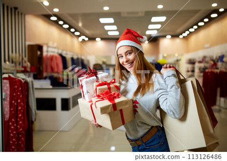 Happy girl shopping gifts in mall on christmas sale. New year holidays shopping idea concept. Smiling woman with colorful paper presents bags and gift boxes wearing christmas hat in store or shop. Happy girl shopping gifts in mall on christmas sale. New year holidays shopping idea concept. Smiling woman with colorful paper presents bags and gift boxes wearing christmas hat in store or shop. 113126486