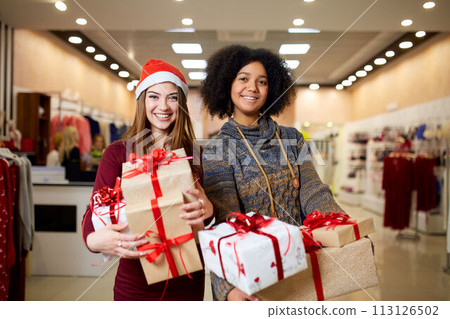 Two mixed race women with gift boxes in hands at store. Multi ethnic girls smiling with presents on christmas new year sale. Caucasian and african american females shopping presents for holidays. 113126502
