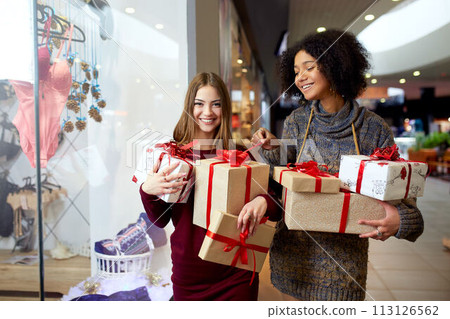 Two mixed race women with gift boxes in hands near storewindow. Multi ethnic girls smiling with presents on christmas new year sale. Caucasian and african american females buy presents for holidays. Two mixed race women with gift boxes in hands near storewindow. Multi ethnic girls smiling with presents on christmas new year sale. Caucasian and african american females buy presents for holidays. 113126562