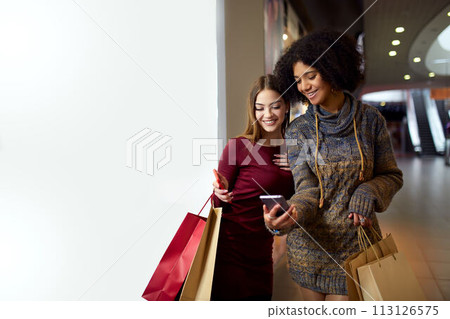 Two happy multiethnic young mixed race woman shopping for lingerie near white shop window copyspace, looking at smartphone screen. African american girl considers underwear with friend at shop front Two happy multiethnic young mixed race woman shopping for lingerie near white shop window copyspace, looking at smartphone screen. African american girl considers underwear with friend at shop front 113126575
