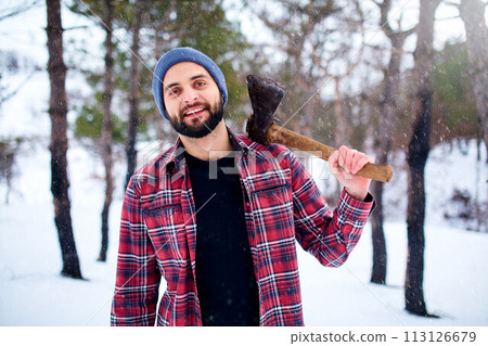 Bearded hipster man in a winter snowy forest with axe on a shoulder. Woodman standing in the forest. Male inspecting trees in woods. Lumberjack woodcutter holding ax wearing plaid red shirt. 113126679