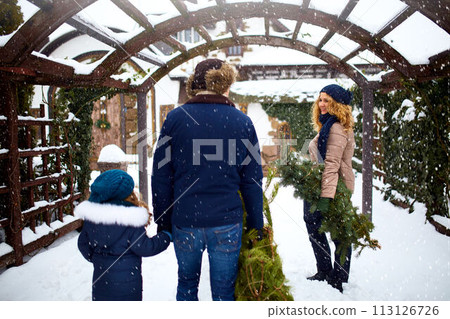 Authentic family carrying Christmas trees and branches to house. Father mother and daughter prepare for New Year and going to decorate their home with spruce and fir tree on celebration. Snowfall. Authentic family carrying Christmas trees and branches to house. Father mother and daughter prepare for New Year and going to decorate their home with spruce and fir tree on celebration. Snowfall. 113126726
