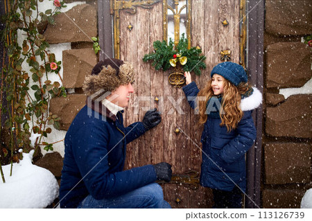 Little girl helps father and hanging Christmas wreath on the door. Cute curly daughter spend time with parents on holidays. New Year home decorations and preparations. Snowy winter outdoors. Little girl helps father and hanging Christmas wreath on the door. Cute curly daughter spend time with parents on holidays. New Year home decorations and preparations. Snowy winter outdoors. 113126739