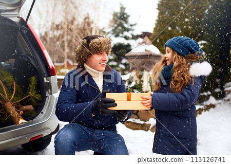 Father presents daughter a gift box on snowy winter day outdoors. Christmas tree in large trunk of family car. Girl helps dad with New Year home decorations and preparations for holidays. Snowfall. 113126741