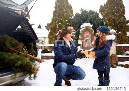 Father presents daughter a gift box on snowy winter day outdoors. Christmas tree in large trunk of family car. Girl helps dad with New Year home decorations and preparations for holidays. Snowfall. Father presents daughter a gift box on snowy winter day outdoors. Christmas tree in large trunk of family car. Girl helps dad with New Year home decorations and preparations for holidays. Snowfall. 113126743