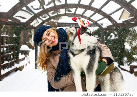 Attractive authentic caucasian woman hugs funny malamute dog wearing santa dear christmas antlers. Curly smiling female having fun with huskie puppy on new year. Pet is best gift for holidays. 113126753