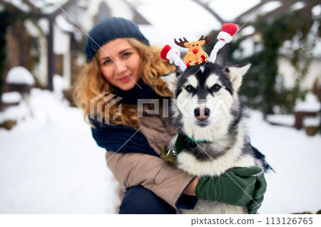 Attractive authentic caucasian woman hugs funny malamute dog wearing santa dear christmas antlers. Curly smiling female having fun with huskie puppy on new year. Pet is best gift for holidays. 113126765
