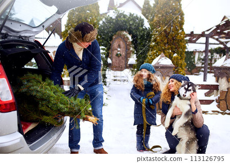 Father brought christmas tree in trunk of SUV car to daughter, mother and dog to decorate home. Family prepares for new year together. Large boot space concept. Snowy winter outdoors Father brought christmas tree in trunk of SUV car to daughter, mother and dog to decorate home. Family prepares for new year together. Large boot space concept. Snowy winter outdoors 113126795