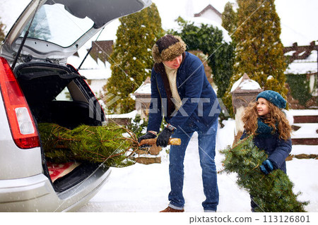 Smiling little girl and father get christmas tree out of car trunk near their house outdoors. Daughter helps dad with new year home decorations and fir-tree on snowy winter. Preparation for holidays. Smiling little girl and father get christmas tree out of car trunk near their house outdoors. Daughter helps dad with new year home decorations and fir-tree on snowy winter. Preparation for holidays. 113126801
