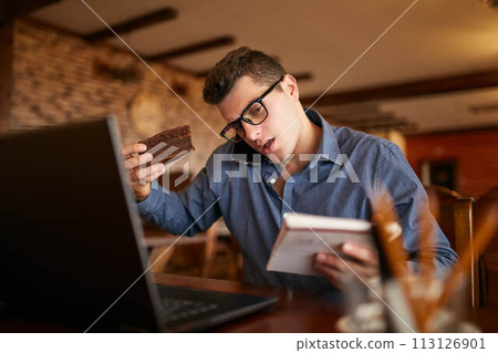 Overworked businessman speaking on cellphone holding it with shoulder, reading writings in notebook, eating a cake and working on laptop simultaneously. Multitasking concept. Busy stylish man. 113126901
