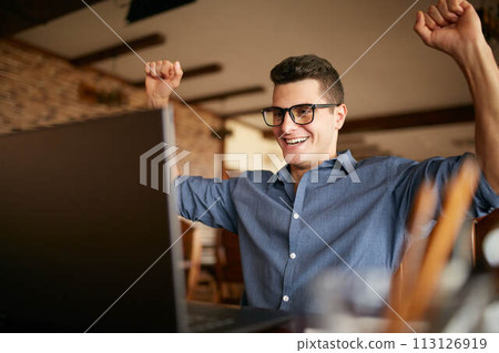 Handsome businessman with laptop having his arms with fists raised, celebrating success. Happy freelancer hipster in glasses finished work on project. Man won a lot of money in lottery prize. 113126919