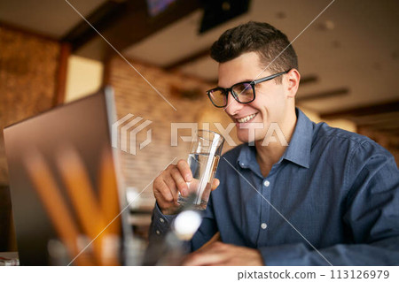 Smiling caucasian man with glass of water in hand works with laptop. Businessman in glasses drinks water for body hydration while working. Attractive designer quench thirst. Healthy lifestyle theme. Smiling caucasian man with glass of water in hand works with laptop. Businessman in glasses drinks water for body hydration while working. Attractive designer quench thirst. Healthy lifestyle theme. 113126979