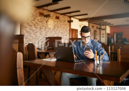 A young man typing a text on mobile modern smartphone. Hipster holding a modern phone and writing a phone message. Smiling young businessman in glasses looking at cellphone with laptop on table. 113127006