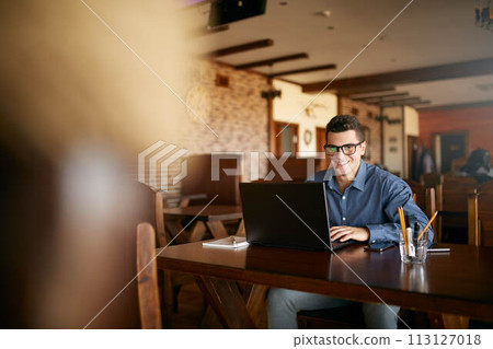 Authentic portrait of young smiling businessman looking at camera with laptop in cafe. Hipster like man in stylish glasses and smart casual shirt doing his work on project. Smart casual wear. Authentic portrait of young smiling businessman looking at camera with laptop in cafe. Hipster like man in stylish glasses and smart casual shirt doing his work on project. Smart casual wear. 113127018