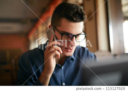 Young freelancer working with laptop and talking on cellphone with client in office. Handsome caucasian businessman in glasses conducts negotiations via phone call. Multitasking business concept. 113127020