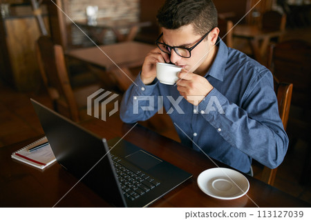 Caucasian businessman using phone while working on laptop and holding cup of coffee in hand. Multitasking concept. Busy freelancer in glasses drinks tea and talking on cellphone in cafe. Smart casual. Caucasian businessman using phone while working on laptop and holding cup of coffee in hand. Multitasking concept. Busy freelancer in glasses drinks tea and talking on cellphone in cafe. Smart casual. 113127039