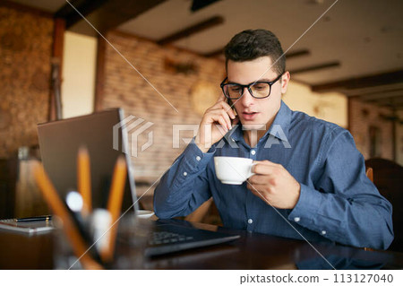 Caucasian businessman using phone while working on laptop and holding cup of coffee in hand. Multitasking concept. Busy freelancer in glasses drinks tea and talking on cellphone in cafe. Smart casual. Caucasian businessman using phone while working on laptop and holding cup of coffee in hand. Multitasking concept. Busy freelancer in glasses drinks tea and talking on cellphone in cafe. Smart casual. 113127040