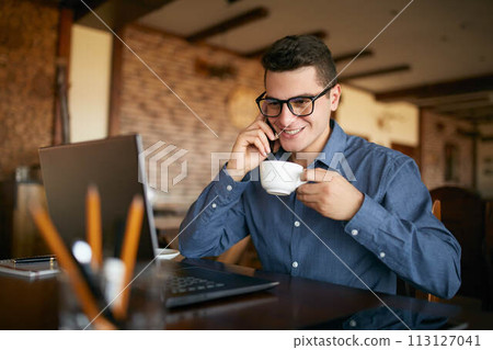 Smiling businessman using phone while working on laptop and holding cup of coffee in hand. Multitasking concept. Busy freelancer in glasses drinks tea and talking on cellphone in cafe. Smart casual. Smiling businessman using phone while working on laptop and holding cup of coffee in hand. Multitasking concept. Busy freelancer in glasses drinks tea and talking on cellphone in cafe. Smart casual. 113127041