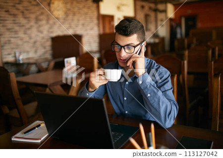 Caucasian businessman using phone while working on laptop and holding cup of coffee in hand. Multitasking concept. Busy freelancer in glasses drinks tea and talking on cellphone in cafe. Smart casual. Caucasian businessman using phone while working on laptop and holding cup of coffee in hand. Multitasking concept. Busy freelancer in glasses drinks tea and talking on cellphone in cafe. Smart casual. 113127042