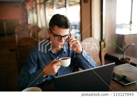 Caucasian businessman using phone while working on laptop and holding cup of coffee in hand. Multitasking concept. Busy freelancer in glasses drinks tea and talking on cellphone in cafe. Smart casual. Caucasian businessman using phone while working on laptop and holding cup of coffee in hand. Multitasking concept. Busy freelancer in glasses drinks tea and talking on cellphone in cafe. Smart casual. 113127043