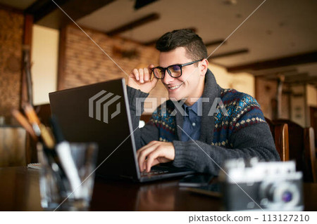 Smiling attractive hipster man in glasses and winter sweater working on laptop in cafe. Freelancer businessman browsing internet at coffeshop. Photographer retouching photos. Vintage camera on table. Smiling attractive hipster man in glasses and winter sweater working on laptop in cafe. Freelancer businessman browsing internet at coffeshop. Photographer retouching photos. Vintage camera on table. 113127210