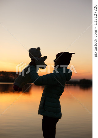 Silhouette of mother holding little baby in her hands on sunset with lake on background. Female with child. Family concept. Mother's day theme. 113127226