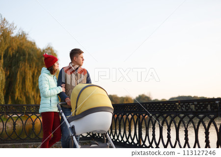A young loving family walks by the lake with a stroller. Smiling parents couple with baby pram in autumn park. Love, parenthood, family, season and people concept. A young loving family walks by the lake with a stroller. Smiling parents couple with baby pram in autumn park. Love, parenthood, family, season and people concept. 113127246