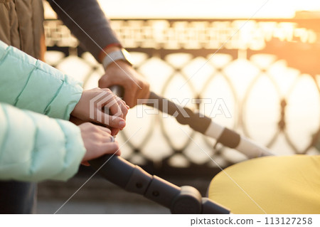 Close up of young loving family hands holding baby pram by the lake. Parents with stroller in autumn park on sunset. Love, parenthood, family concept. 113127258
