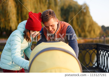 A young loving family walks by the lake with a stroller. Smiling parents couple with baby pram in autumn park look into cariage. Love, parenthood, family, season and people concept. 113127281