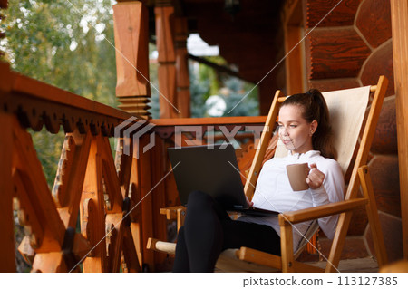 Young attractive business woman is working on the laptop at mountain resort with a cup of tea and cookie. Female relaxes on deck chair with notebook and coffee. Young attractive business woman is working on the laptop at mountain resort with a cup of tea and cookie. Female relaxes on deck chair with notebook and coffee. 113127385