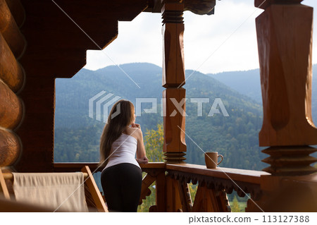 Woman leaning on wooden handrail and enjoys and relaxes beautiful mountain scenic. Young female on terrace leaning on handrail with a cup of tea or coffee 113127388
