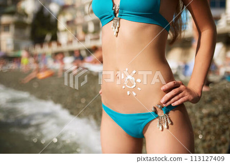 Water resistant sunscreen lotion drawn on tanned woman belly skin as sun shape. Woman wearing bikini swimsuit sunbathing on the beach standing in the sea water. Suntan cream on stomach. 113127409