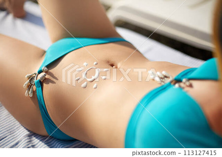 Sunscreen lotion drawn on tanned woman belly skin as sun shape. Suntan cream applied on body. Suncream on stomach. Woman wearing bikini swimsuit sunbathing on the beach. Skin care health concept. Sunscreen lotion drawn on tanned woman belly skin as sun shape. Suntan cream applied on body. Suncream on stomach. Woman wearing bikini swimsuit sunbathing on the beach. Skin care health concept. 113127415