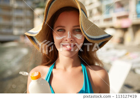Suntan lotion. Woman applying sunscreen solar cream on face. Beautiful happy cute girl puts suntan cream from plastic container bottle on her nose and cheecks. Female in straw hat and bikini on beach. Suntan lotion. Woman applying sunscreen solar cream on face. Beautiful happy cute girl puts suntan cream from plastic container bottle on her nose and cheecks. Female in straw hat and bikini on beach. 113127419
