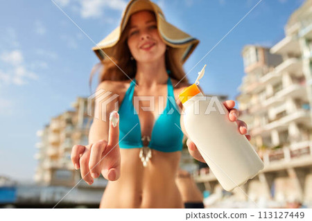 Closeup of smiling caucasian woman showing sunscreen oil bottle and using sun protection cream smear on finger. Pretty tanned girl wearing bikini swimsuit, straw hat and sunglasses on beach near sea. Closeup of smiling caucasian woman showing sunscreen oil bottle and using sun protection cream smear on finger. Pretty tanned girl wearing bikini swimsuit, straw hat and sunglasses on beach near sea. 113127449