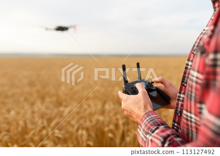 Farmer holds remote controller with his hands while quadcopter is flying on background. Drone hovers behind the agronomist in wheat field. Agricultural new technologies and innovations. Back view Farmer holds remote controller with his hands while quadcopter is flying on background. Drone hovers behind the agronomist in wheat field. Agricultural new technologies and innovations. Back view 113127492