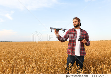 Bearded hipster man shows small compact drone and holds remote controller in his hand. Farmer agronomist looks at quadcopter before the launch in a wheat field Bearded hipster man shows small compact drone and holds remote controller in his hand. Farmer agronomist looks at quadcopter before the launch in a wheat field 113127508