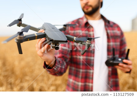 Bearded hipster man shows small compact drone and holds remote controller in his hand. Farmer agronomist looks at quadcopter before the launch in a wheat field 113127509