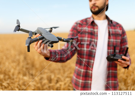 Bearded hipster man shows small compact drone and holds remote controller in his hand. Farmer agronomist looks at quadcopter before the launch in a wheat field Bearded hipster man shows small compact drone and holds remote controller in his hand. Farmer agronomist looks at quadcopter before the launch in a wheat field 113127511