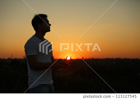 Silhouette of a man piloting drone in the air with a remote controller in his hands on sunset. Pilot takes aerial photos and videos with quadcopter from above 113127545