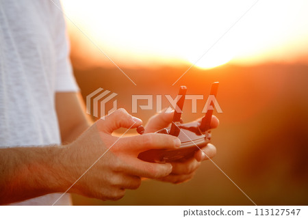 Man holds a drone remote controller in his hands. Close-up of quadrocopter RC during flight. Pilot takes aerial photos and videos with quad on sunset 113127547