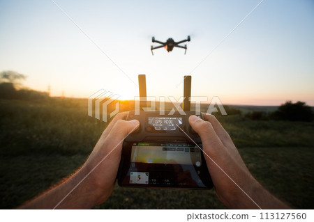 Point of view shot of man holding remote controller with his hands and taking aerial photo video. Quadcopter is flying on background. POV - drone hovers in front of the pilot on suset 113127560