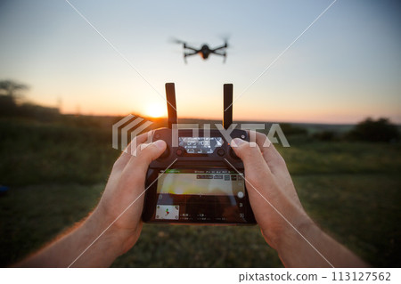 Point of view shot of man holding remote controller with his hands and taking aerial photo video. Quadcopter is flying on background. POV - drone hovers in front of the pilot on suset 113127562